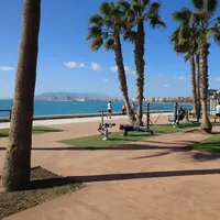 Parque de ejercicios del Paseo Marítimo — Small plaza featuring outdoor gym equipment, benches & a pavilion with palm trees & sea views.